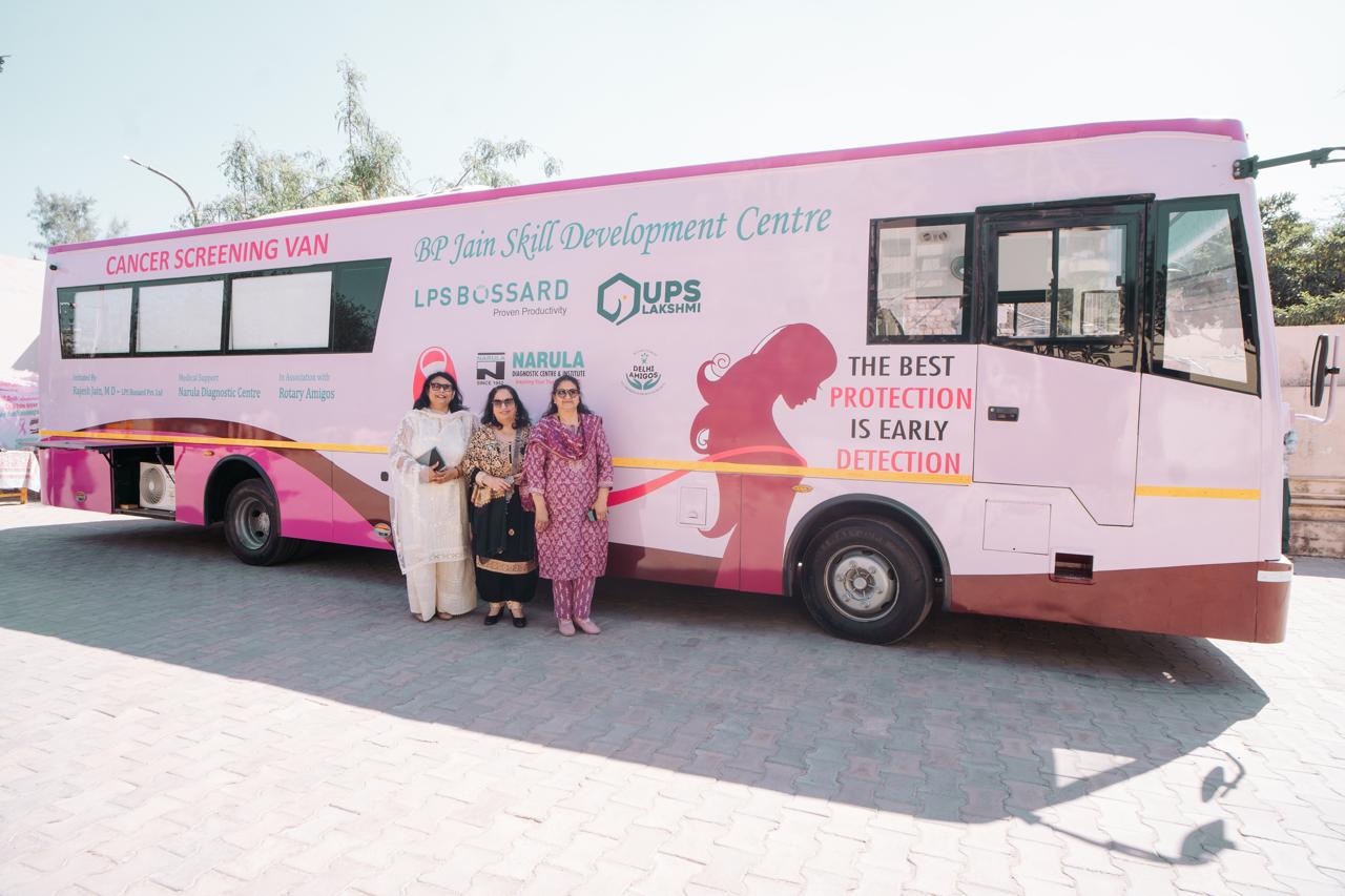The image shows a large mobile cancer screening van painted in white and pink, labeled “Cancer Screening Van” and “BP Jain Skill Development Centre.” The van displays messages about the importance of early detection in preventing cancer. Three women are standing in front of the van, posing for the photograph. The vehicle features branding from supporting organizations such as LPS Bossard, UPS Lakshmi, and Narula Diagnostics Centre. The slogan on the van reads, “The best protection is early detection,” highlighting the awareness and preventive healthcare focus of the initiative.