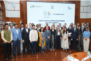 A group of approximately 30 individuals, primarily men and women dressed in business casual attire and traditional Indian clothing, stand together in a large room with wooden walls and white ceilings. A prominent banner behind them reads "India-France AI Policy Roundtable - From Action to Impact."