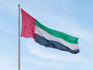 The flag of the United Arab Emirates (UAE) flying against a blue sky with light clouds. The flag features horizontal stripes of green, white, and black, with a vertical red stripe on the hoist side.