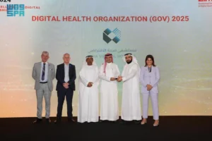  A group of six people, four men and two women, stand on a stage in front of a large screen displaying the words "DIGITAL HEALTH ORGANIZATION (GOV) 2025" and the logo for "Seha Virtual Hospital." Three men in the center are wearing traditional white thobes and keffiyehs, one of whom is holding a clear award. Two men on the left are in suits, and a woman on the right is in a light purple pantsuit.