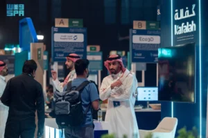 Several men in a professional setting, some wearing traditional Saudi attire, are gathered around a counter with informational displays in the background.