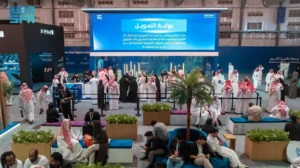 A wide shot of a bustling indoor forum with many attendees, primarily men in traditional Middle Eastern attire, and some women in abayas. In the center, a large screen displays Arabic text with the words "بوابة التمويل" (The Funding Gate) and "SME Bank." There are several booths and seating areas with plants, and people are networking, sitting, and looking at their phones.