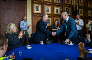 Two men shaking hands across a blue round table in a room with several other people, some clapping. One man wears a dark suit, the other a blue sweater.