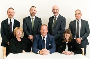  A group of seven business professionals, four men standing and three people seated, looking towards the camera with friendly expressions. The standing men are wearing dark suits and ties, while the seated individuals include two women in dark blazers and a man in a blue suit and tie. They appear to be in a professional setting with a plain white background.