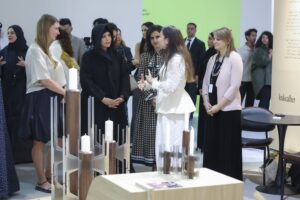 Latifa bint Mohammed, dressed in a black abaya, stands among a group of people at what appears to be an exhibition. She is looking towards a woman in a white lace top and white trousers who is gesturing with her hands while speaking. To the right, another woman in a light pink blazer and black skirt looks on. There are various modern furniture pieces and design elements visible, including white candles in wooden holders in the foreground.