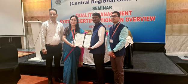 Four people standing on a stage, two holding up certificates, in front of a banner for a "Quality Management System Certification" seminar.