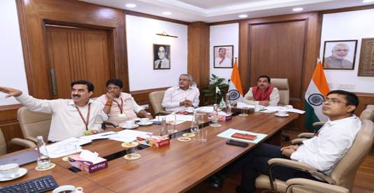 A group of Indian government officials in a meeting room, some seated around a large wooden table with drinks and papers, and one man standing and gesturing. Two Indian flags are visible, and portraits of other officials hang on the walls.