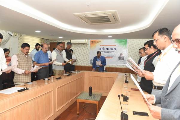  A group of people, mostly men, in an office setting, standing around a large U-shaped wooden table, holding papers and seemingly singing or reciting. There's a banner in the background with "Vande Mataram" written in Hindi and an Indian flag.