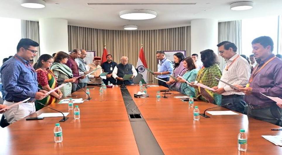  A group of people, appearing to be officials, are gathered in a modern conference room. Many of them are standing and holding papers, seemingly participating in an oath-taking ceremony or a similar official event. Two Indian flags are visible in the background, flanking a central figure seated at the head of a long table.
