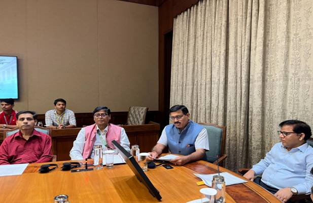 A group of five men are seated around a large wooden conference table, with one man speaking from the center.