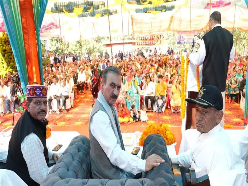 Chief Minister of Himachal Pradesh, with other dignitaries, seated on a stage at a large public gathering or political rally under a festive, colorful canopy, as a speaker addresses the audience.