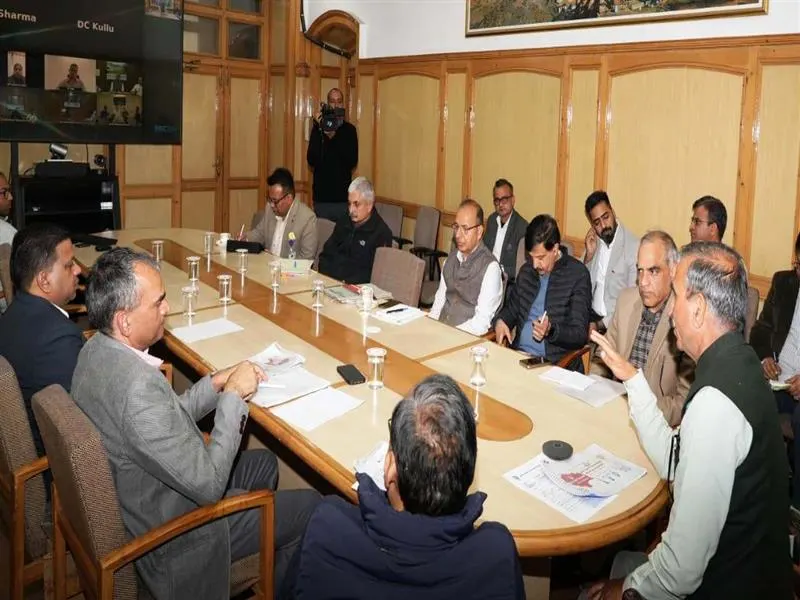 A high-level meeting in a wood-paneled conference room with multiple government officials seated around a large table, engaging in a discussion, while a video conference with remote participants (labeled DC Kullu) is displayed on a screen.