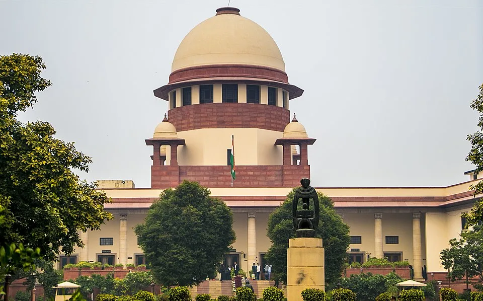 The Supreme Court of India building in New Delhi, featuring its large, central dome and red sandstone façade, framed by green trees and a foreground statue.