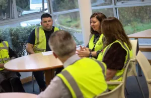 A group of five people wearing high-visibility vests are seated around a circular table in a meeting or breakroom setting.