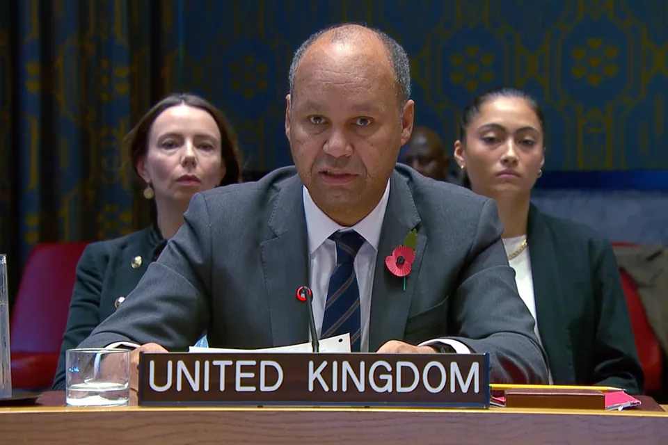 A man in a suit speaking at a "UNITED KINGDOM" desk, flanked by two women, one with dark hair and another with light hair. He wears a poppy pin on his lapel.