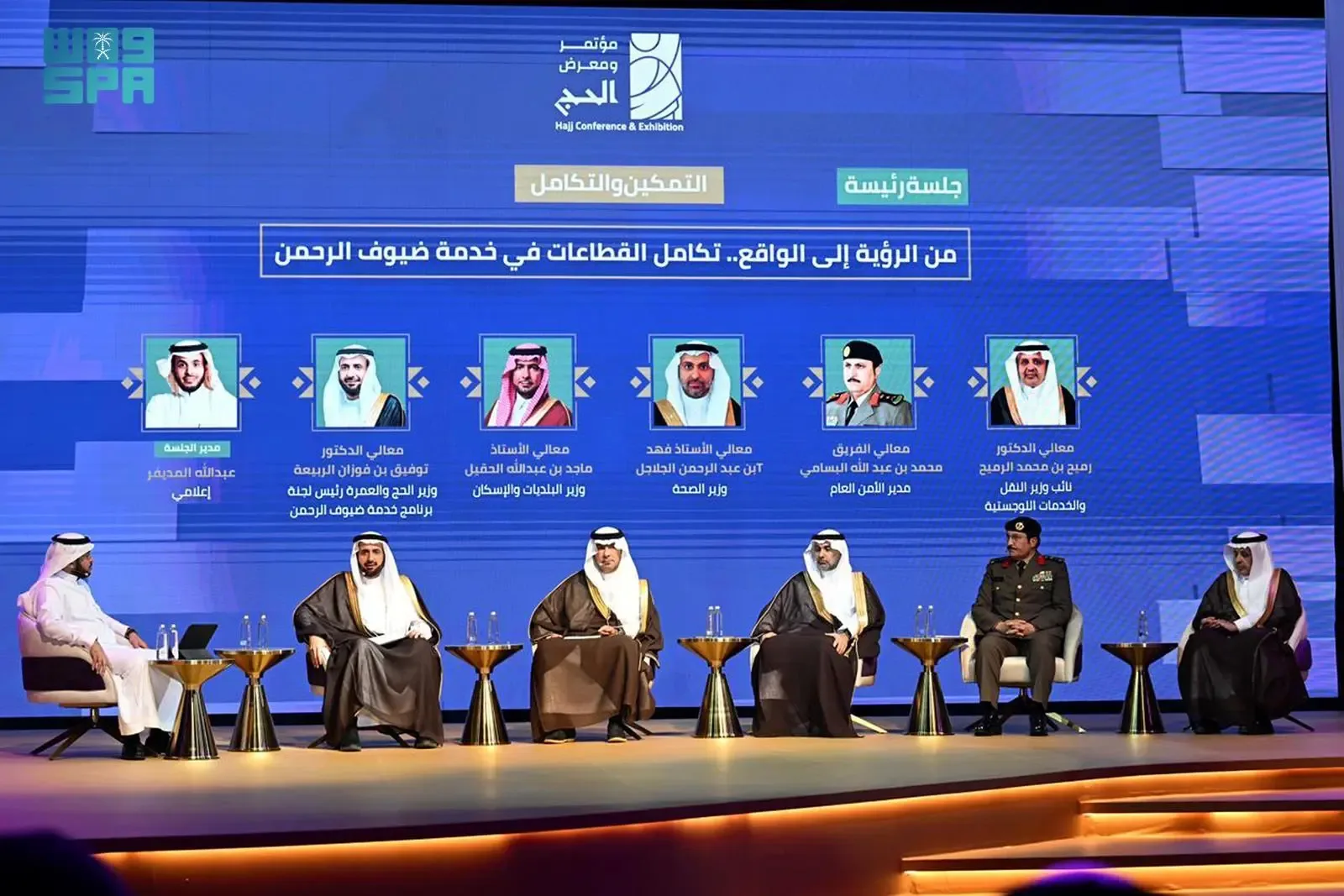 A panel of seven men in traditional Middle Eastern attire and military uniform seated on a stage, with a large blue screen displaying text and images behind them.