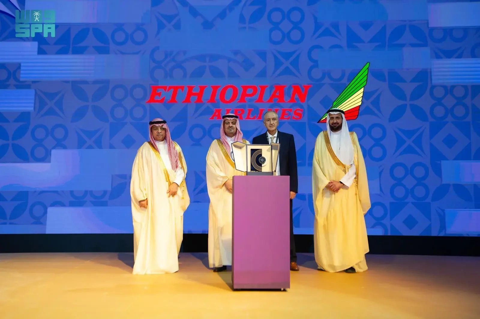 Four men, three in traditional Saudi attire and one in a dark suit, standing on a stage with a blue background displaying the "ETHIOPIAN AIRLINES" logo. A podium is in the center foreground.