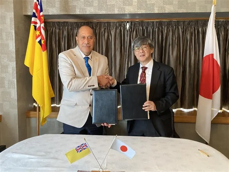 Two men, one in a light suit and the other in a dark suit, shake hands across a table with two flags in front of them, one representing Niue and the other Japan.