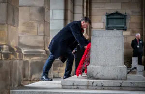 A man in a dark coat laying a red poppy wreath at the base of a stone monument.