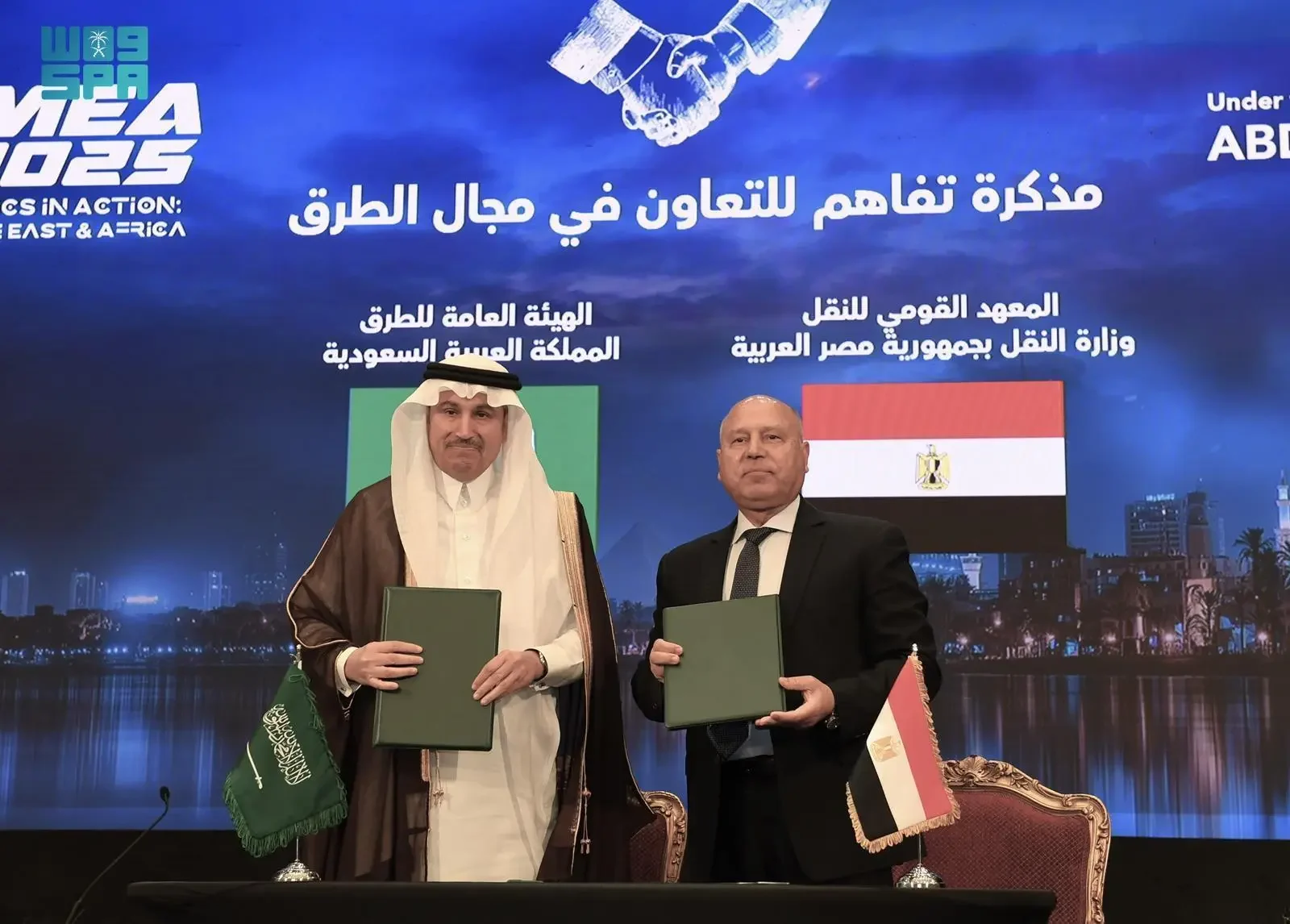 Two men, one in traditional Saudi attire and another in a suit, shake hands after signing documents, with Saudi Arabian and Egyptian flags visible, symbolizing a collaboration agreement.