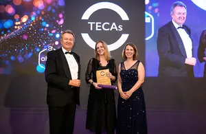 Three individuals on a stage, two men and one woman, presenting an award at the TECAs awards ceremony. The woman in the center is holding the award.
