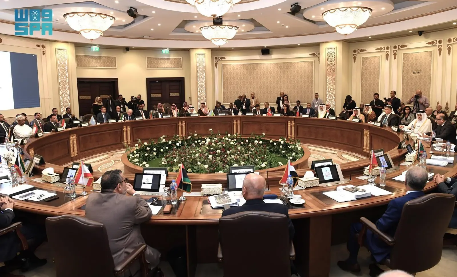 Officials seated around a large circular conference table with a floral centerpiece, participating in a meeting.