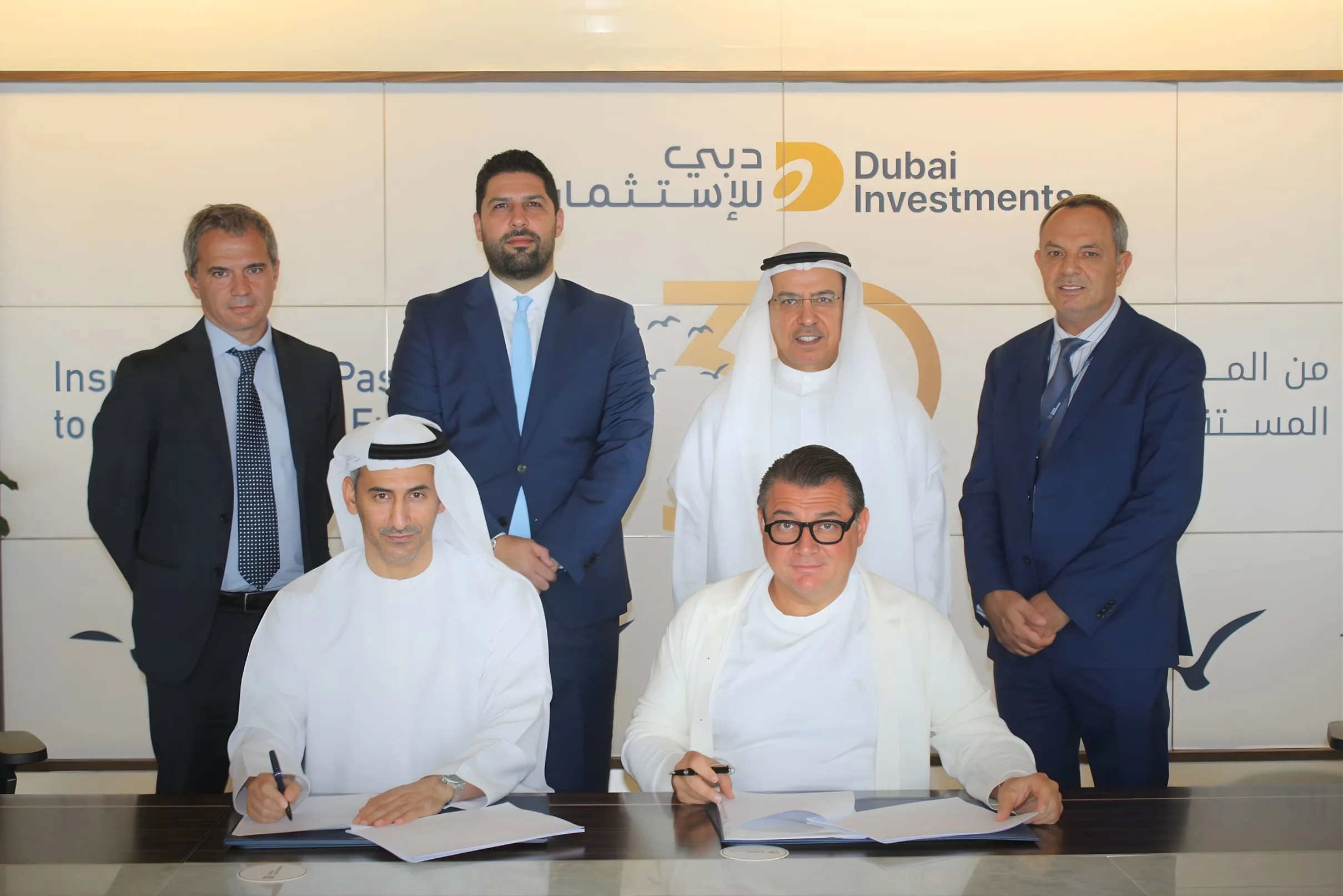 Six men in business attire, some in traditional Emirati dress, are gathered around a conference table. Two men in the foreground are signing documents, while four others stand behind them. A backdrop with the "Dubai Investments" logo is visible.