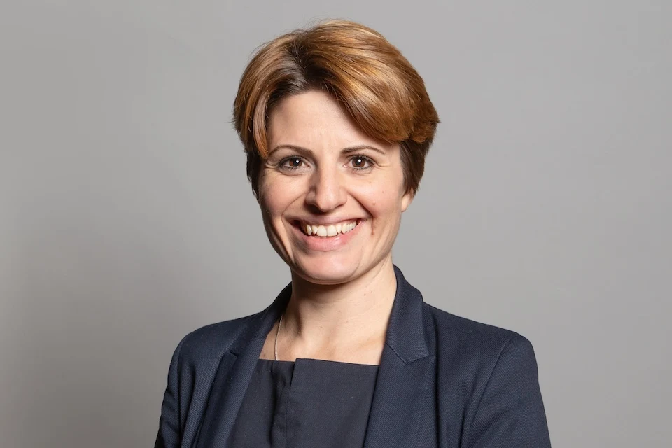 Official portrait of Chloe Smith, a British politician with short brown hair, smiling and wearing a dark blue suit jacket over a dark top.