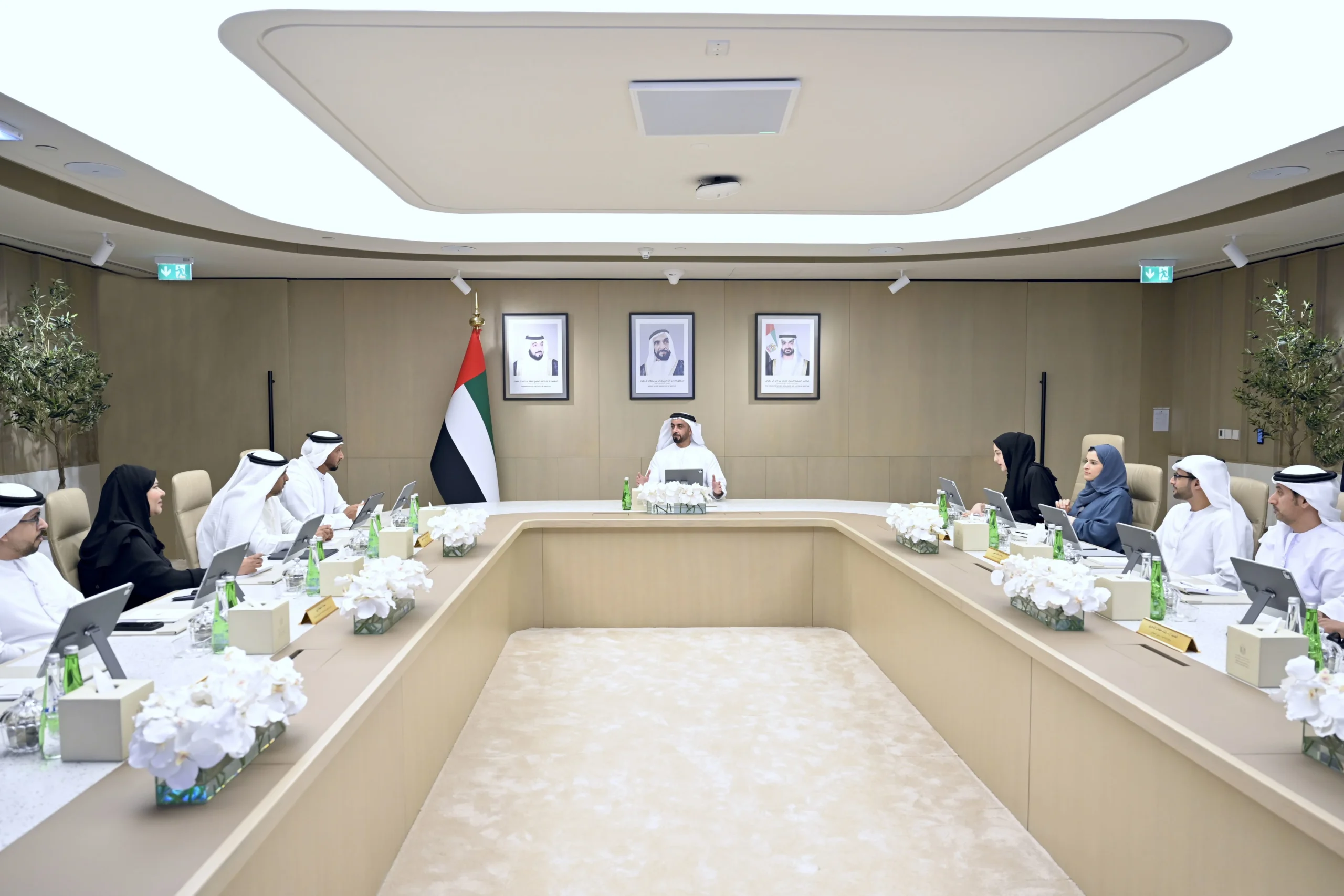 A high-angle shot captures a formal meeting in a spacious conference room, with attendees in traditional attire seated around a large U-shaped table. The room features light wood paneling, recessed lighting, and a flag of the United Arab Emirates. Portraits of leaders adorn the wall behind the central figure.