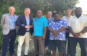 Two men in suits shaking hands with a man in a blue patterned shirt, with four other people standing behind them outdoors.