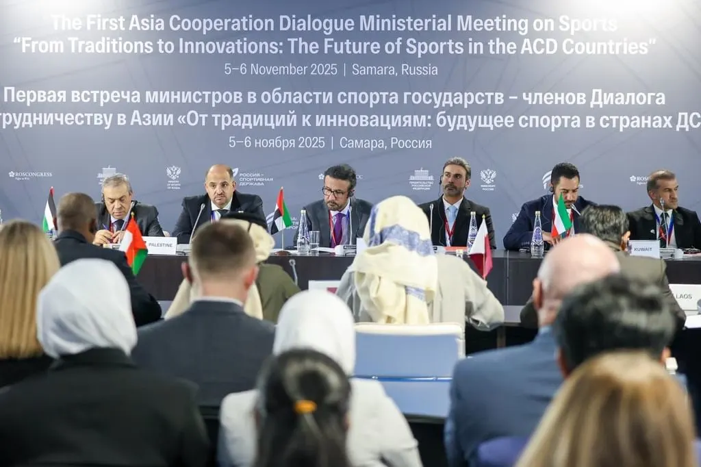 A panel of men in suits, possibly delegates, are seated at a long table with microphones and country flags, while an audience faces them with their backs to the camera. A large screen behind the panel displays text about "The First Asia Cooperation Dialogue Ministerial Meeting on Sports" in English and Russian, with "5-6 November 2025 Samara, Russia" also visible.