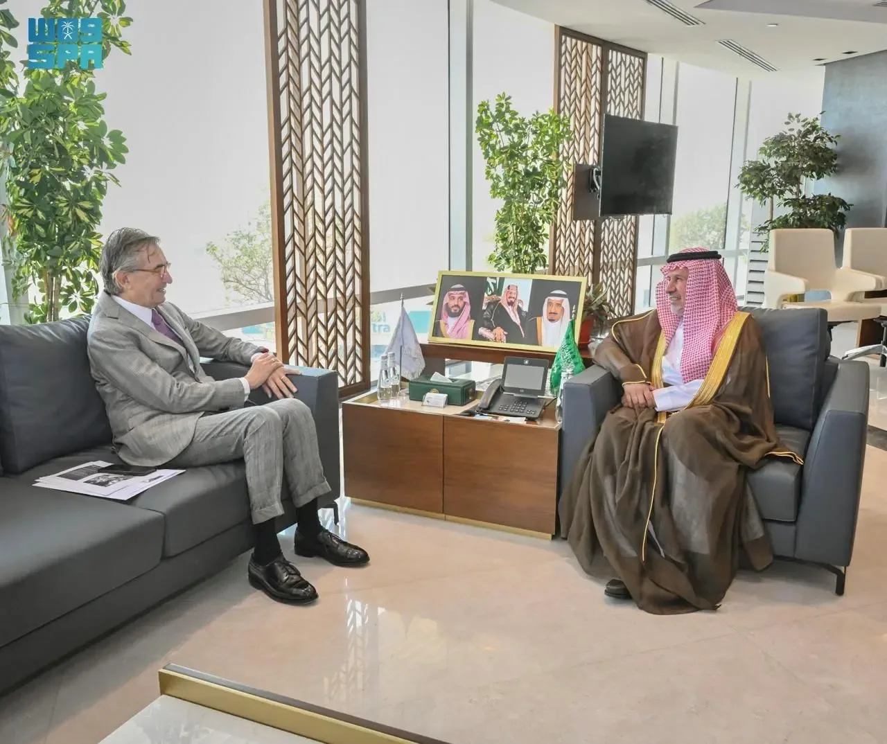 Two men, one in a suit and the other in traditional Saudi attire, are seated on separate sofas in a modern office setting with large windows. There are framed pictures on a side table between them.