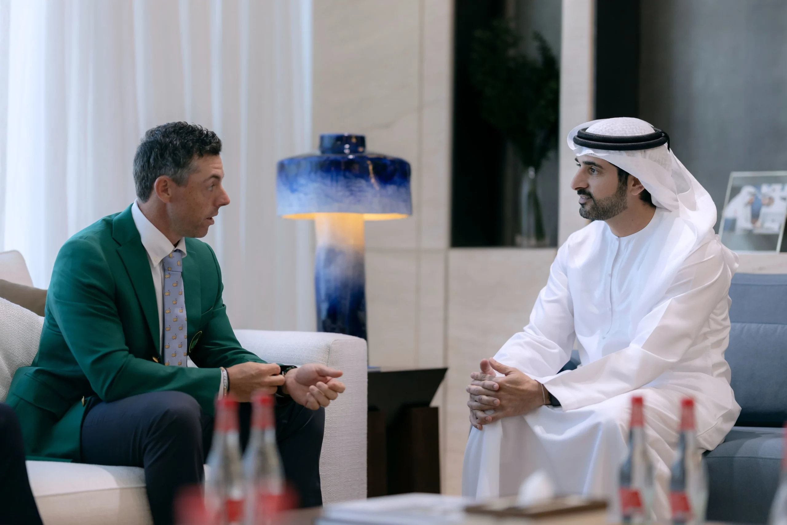 Two men, one in a green suit and the other in traditional white Arab attire, are seated and engaged in conversation indoors. The man on the left has grey hair and is wearing a green blazer over a light blue shirt and patterned tie. The man on the right is wearing a white thobe and ghutra with an agal, and has a beard. They are in a modern room with a blue and white lamp in the background.