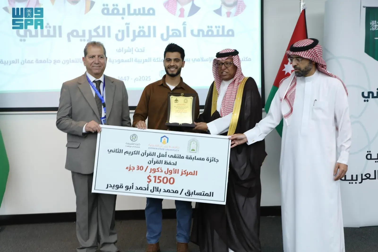 Four men, three in traditional Middle Eastern attire and one in a suit, stand on a stage. One man in traditional attire hands a certificate and a plaque to a younger man in a brown shirt and jeans. The certificate displays Arabic text and "Amman Arab University."
