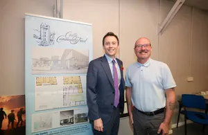 Two men, one in a suit and the other in a polo shirt, standing next to a display board featuring architectural plans and renderings.