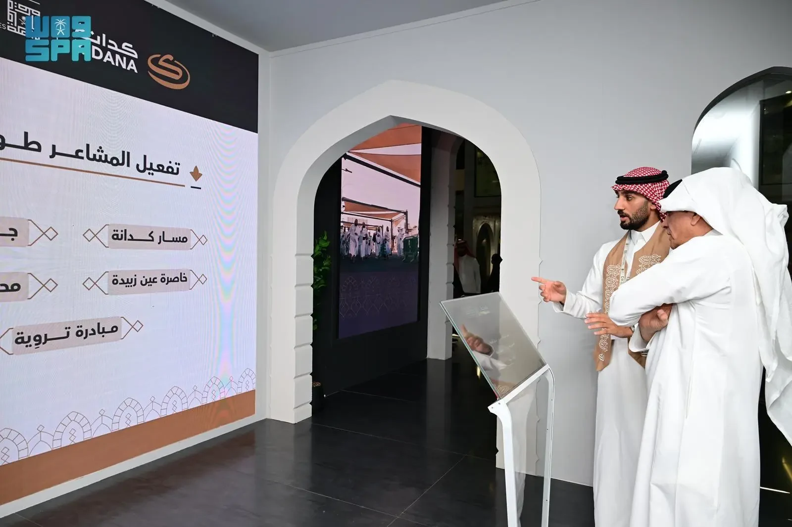 Two men in traditional Saudi attire examining a digital display with Arabic text and graphics, one pointing at the screen, inside a modern exhibition space.