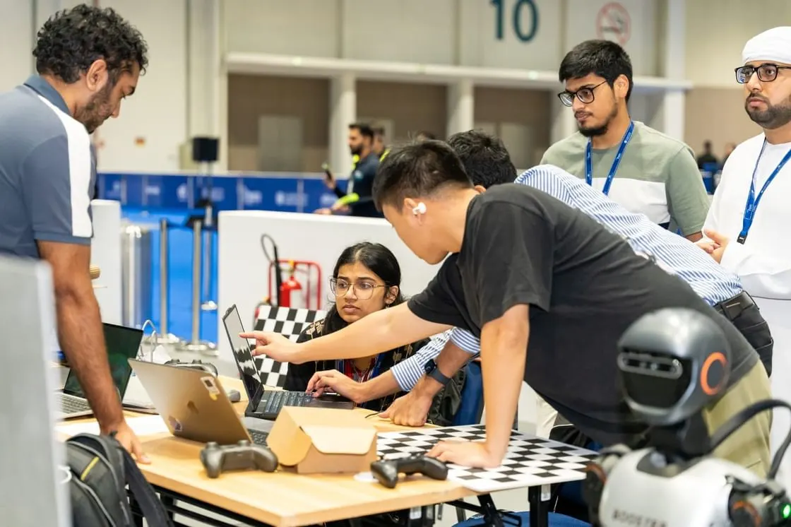 A group of students and mentors collaboratively working on a robotics project with laptops, game controllers, and a robot on a table with a checkerboard pattern.