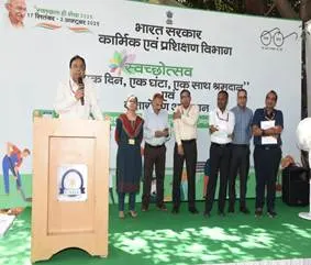  A man speaks at a podium with a microphone, addressing a group of five men and one woman standing in a line on a grassy area in front of a large white banner with Hindi text and a "Swachhta" (cleanliness) logo.
