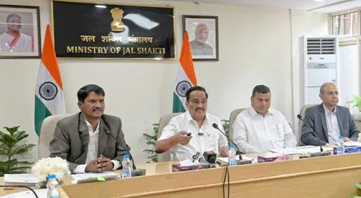 Four men, likely officials, are seated at a long conference table during a meeting. The background features the Ministry of Jal Shakti sign, Indian flags, and framed portraits on the wall.