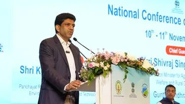 A man in a suit speaking at a podium with microphones, a floral arrangement, and a "National Conference" banner in the background.