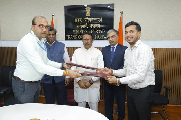 Two men in an office setting shake hands across a desk, exchanging documents, with three other men observing in the background. A sign for the "Ministry of Road Transport and Highways" is visible behind them.
