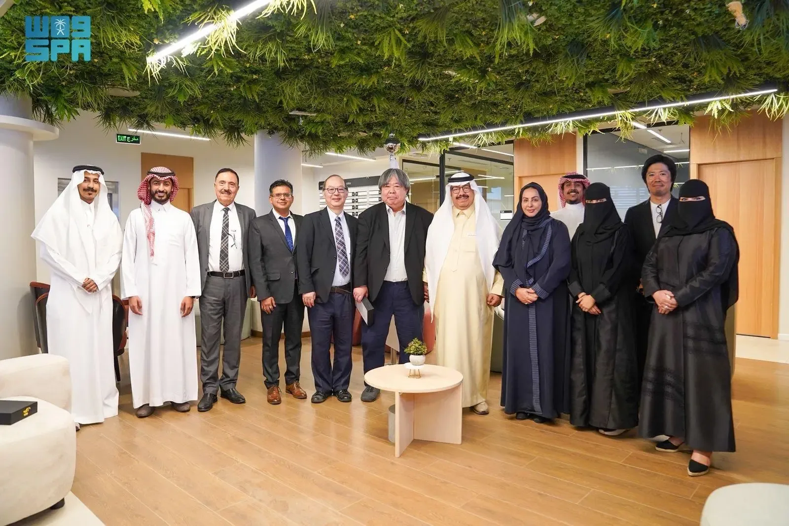A diverse group of men and women, some in traditional Saudi attire and others in business suits, pose for a photo indoors, standing on a wooden floor under a ceiling decorated with greenery.