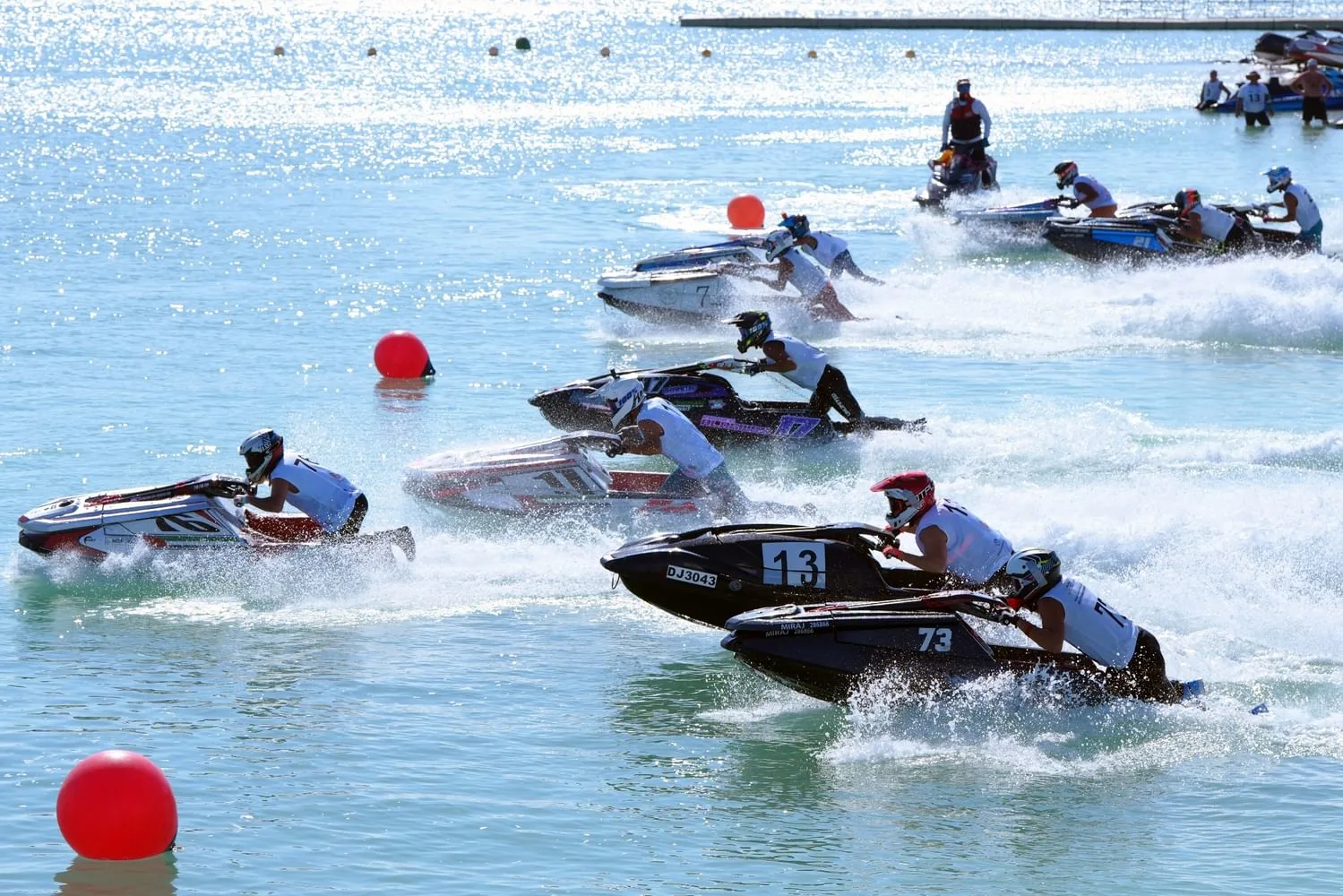 A high-angle shot captures multiple jet skiers racing on bright blue water under sunlight, with red buoys marking the course.