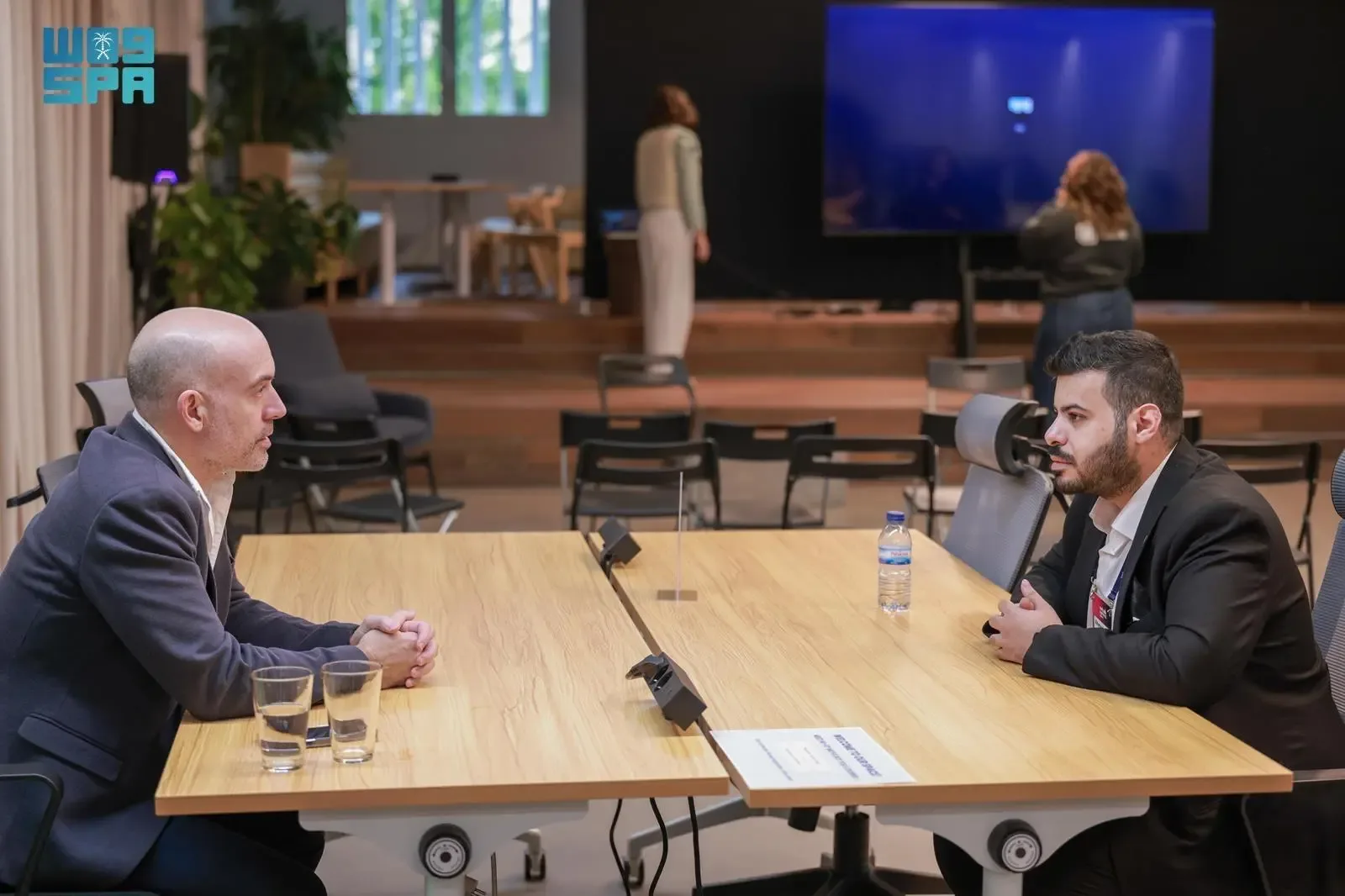 Two men in business attire sit opposite each other at a long wooden table, engaged in conversation. A bottle of water and two glasses are on the table. In the background, other people are visible, and a large screen displays an image or presentation.