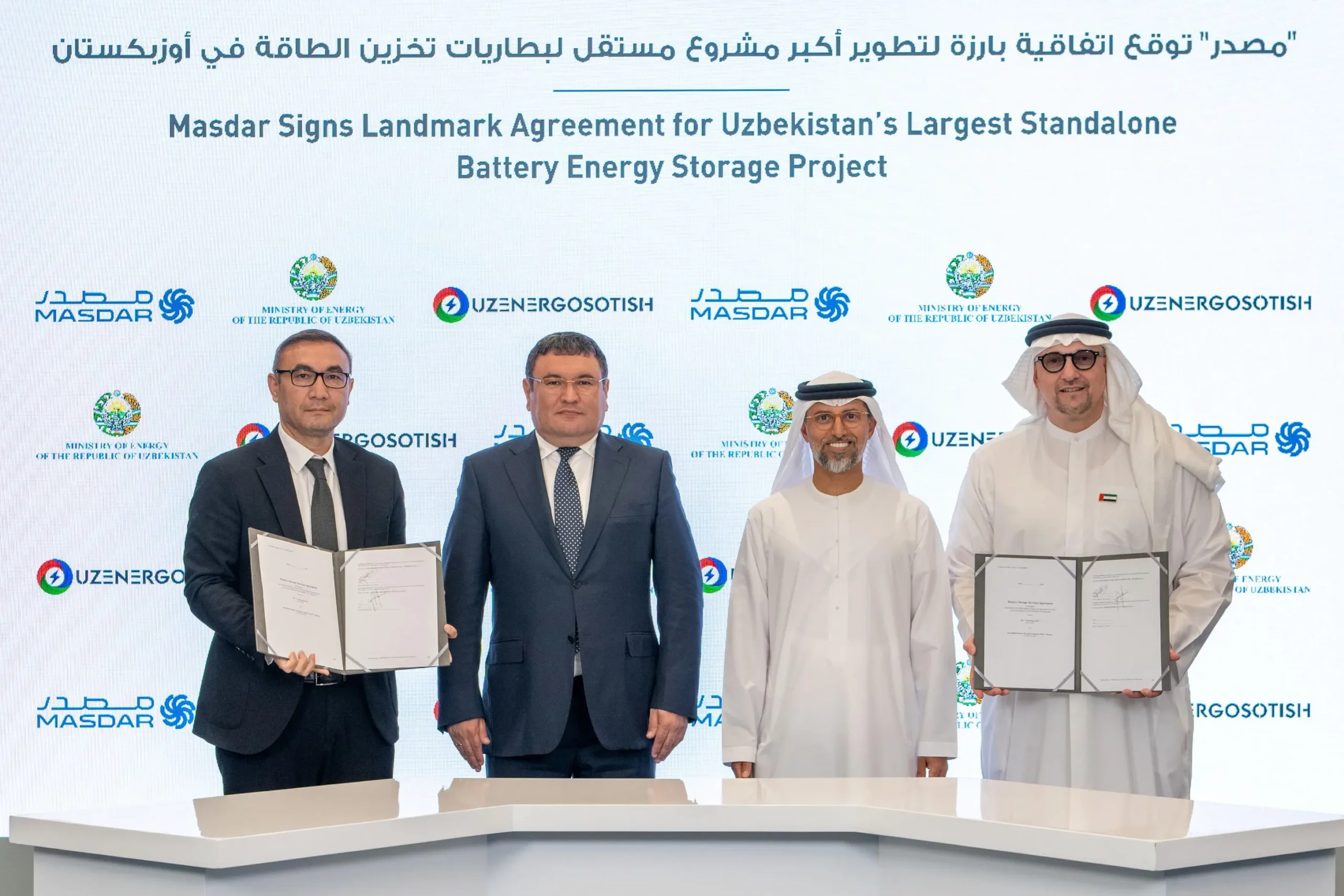 Four men in business attire and traditional wear shaking hands at a signing ceremony, with a backdrop displaying "Masdar Signs Landmark Agreement for Uzbekistan's Largest Standalone Battery Energy Storage Project."