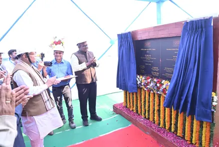 Men in hard hats and traditional Indian attire participate in an unveiling ceremony, with one man holding a plaque while others clap. A draped sign board with text and floral garlands is visible on the right.