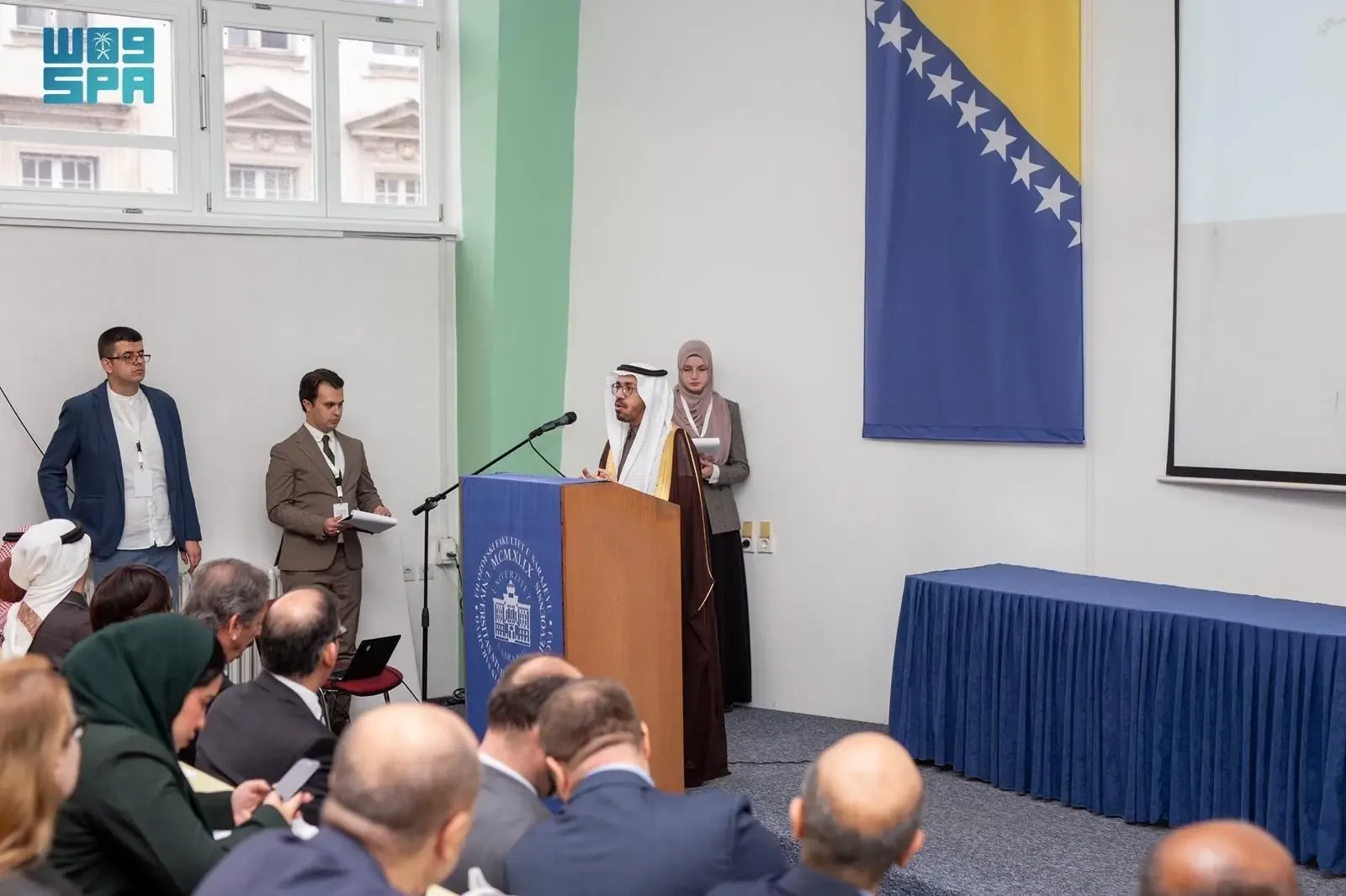 A man in traditional Middle Eastern attire speaks at a podium with a microphone, addressing an audience seated in front of him. A Bosnian flag hangs on the wall behind him, and other individuals are present on the stage.