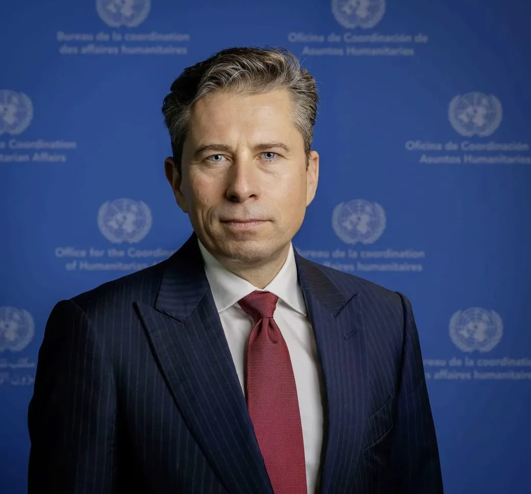 A headshot of a man with short, styled dark hair and blue eyes, wearing a dark pinstripe suit and a red tie, standing in front of a blue background with the United Nations logo and text repeated.