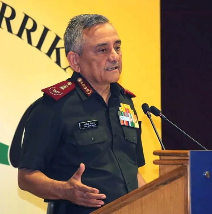 An Indian Army general in uniform speaking at a podium with microphones, a yellow and green banner in the background.