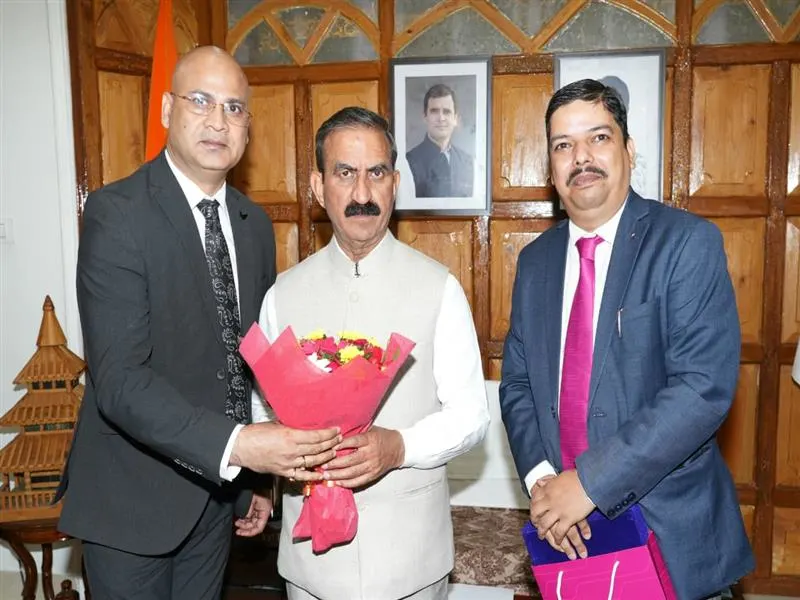 Himachal Pradesh Chief Minister Sukhvinder Singh Sukhu, wearing a light-colored Nehru jacket, receives a bouquet of flowers from a man in a dark suit, while another man in a blue suit stands beside them in an indoor official setting.